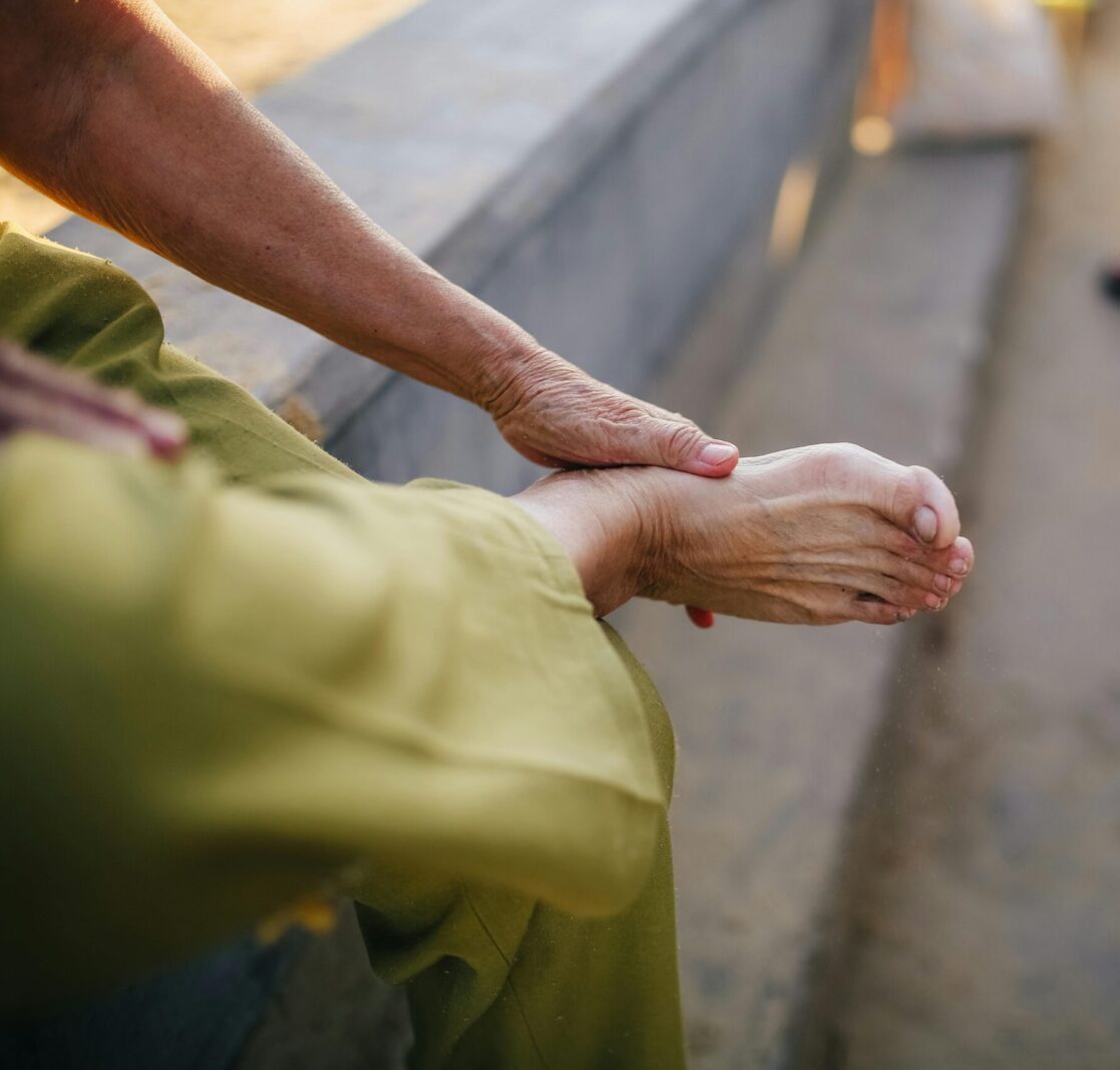 An elderly person sitting outdoors massaging their foot for relief.
